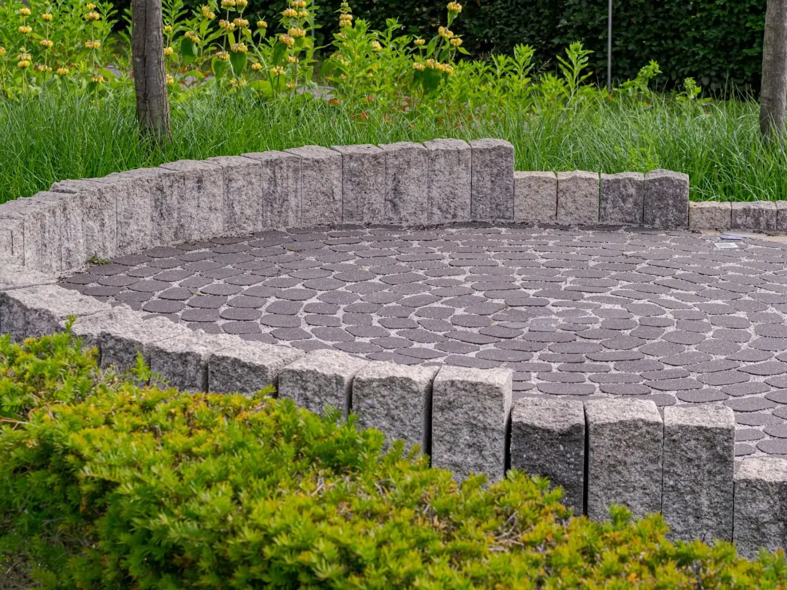 Circular paver patio with a low stone wall and greenery by MCG Top Masonry